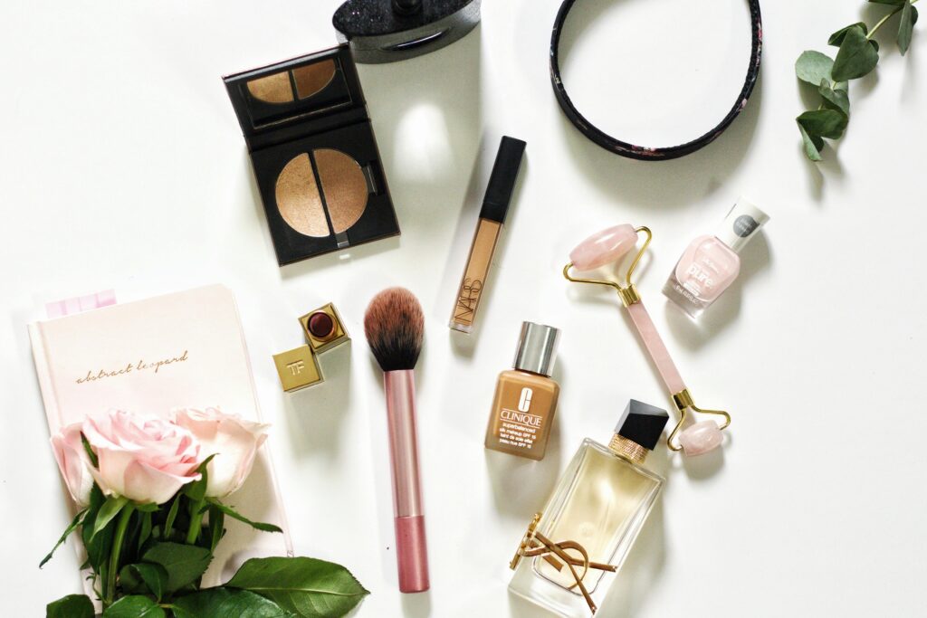 "Beauty and Makeup Products Flat Lay Flat lay of makeup and beauty products, including a foundation, lipstick, concealer, compact, perfume, makeup brush, jade roller, nail polish, notebook, and pink roses on a white background